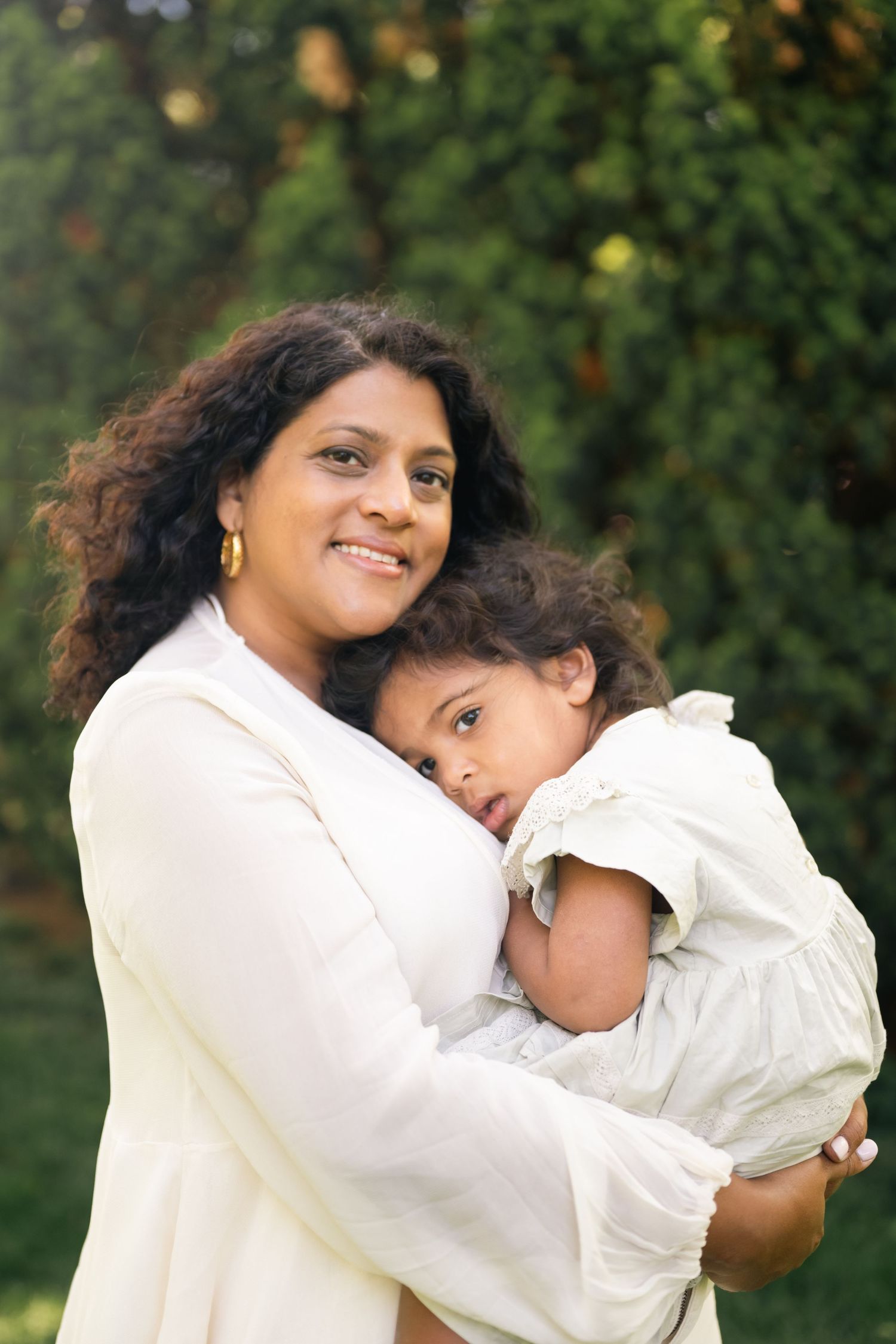 A smiling mother with curly hair holds her young daughter close in a lush green outdoor setting.