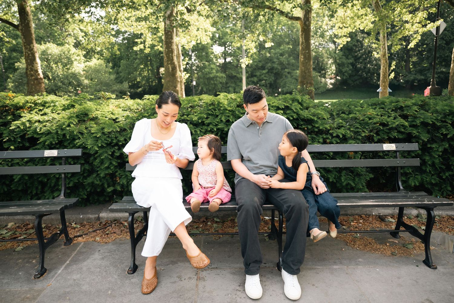 Family of four sitting on a park bench surrounded by lush greenery, enjoying a sunny day together outdoors.