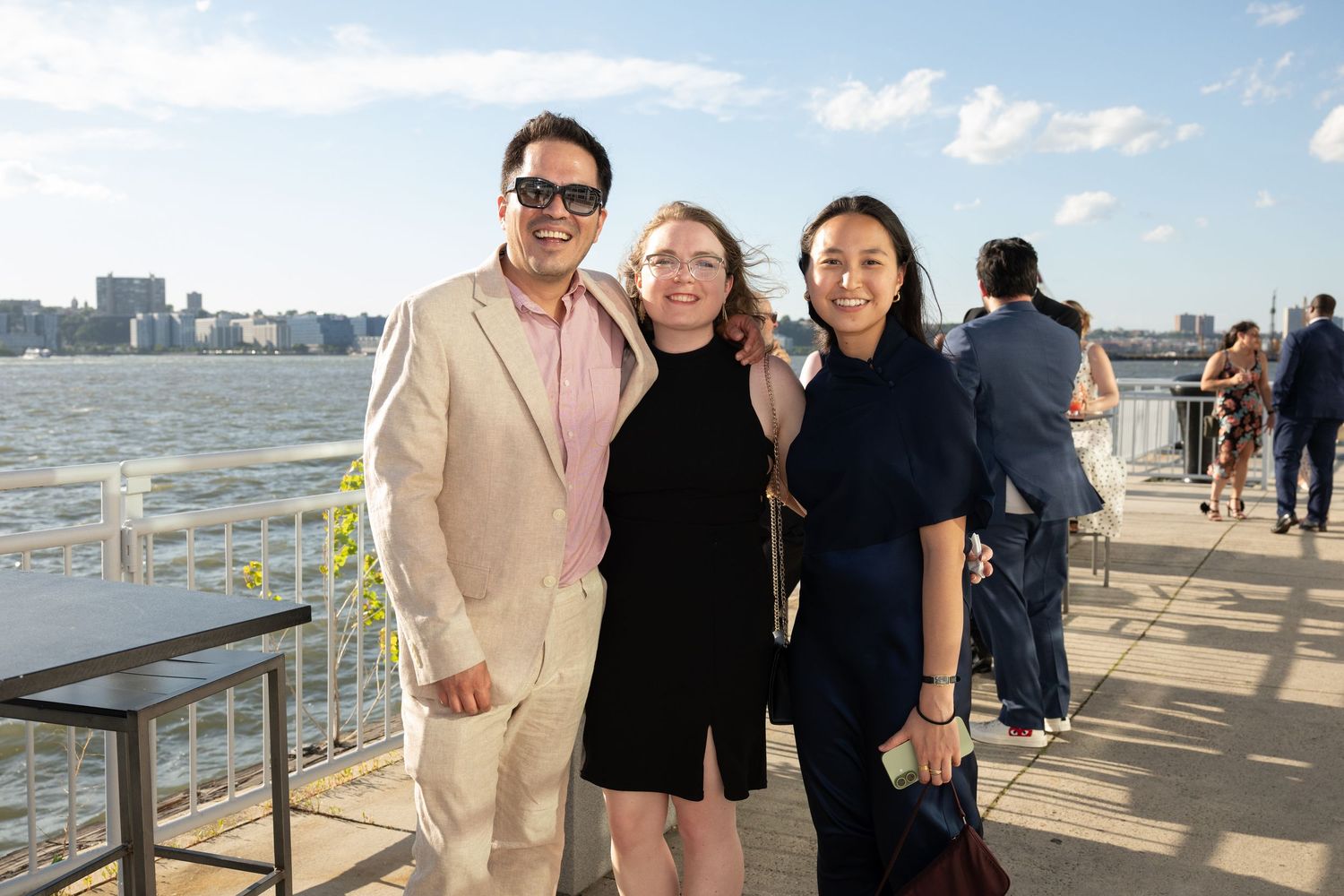 Three guests smiling together on a waterfront pier at an outdoor evening event with city skyline views.