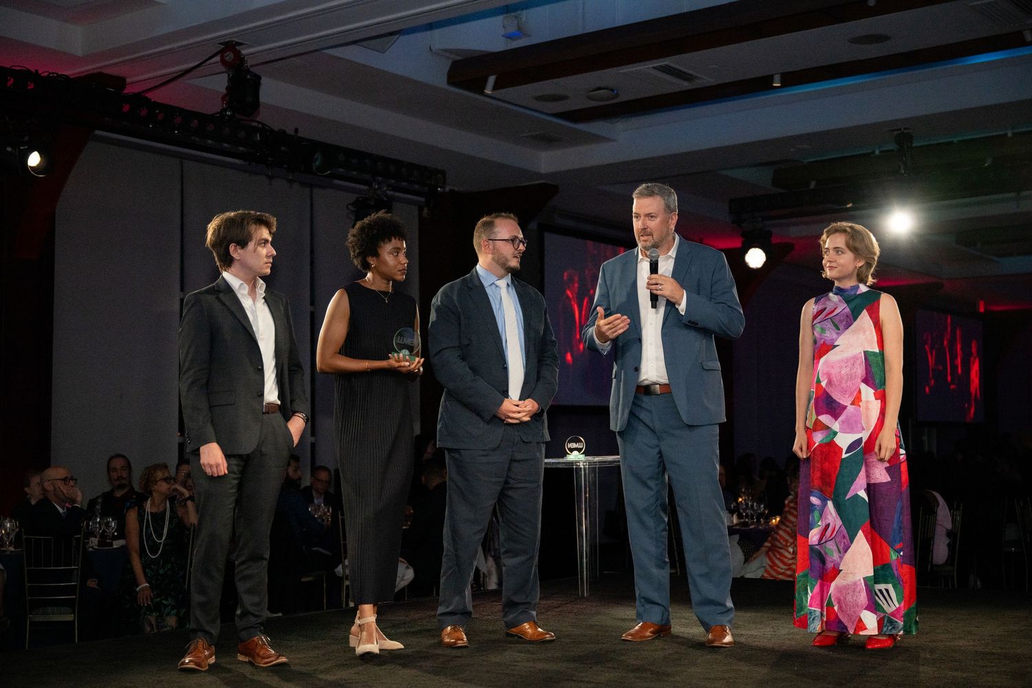 Five people on a stage at an awards ceremony, one speaking into a microphone at an indoor event.