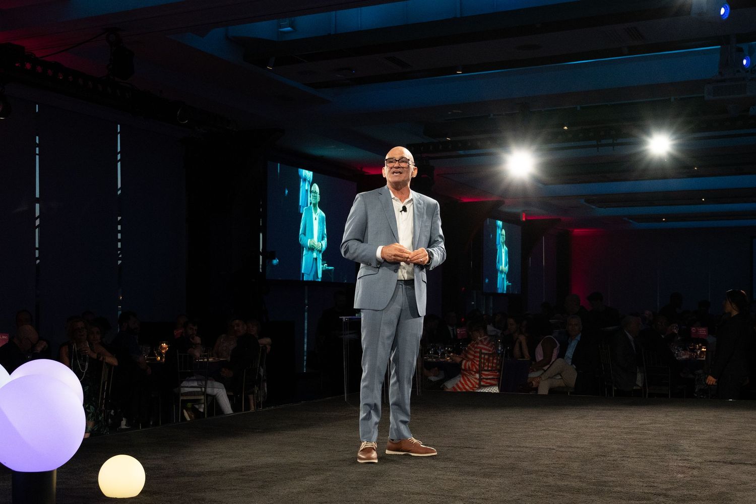 Speaker presenting on stage at a corporate event in a gray suit with audience seated in background.