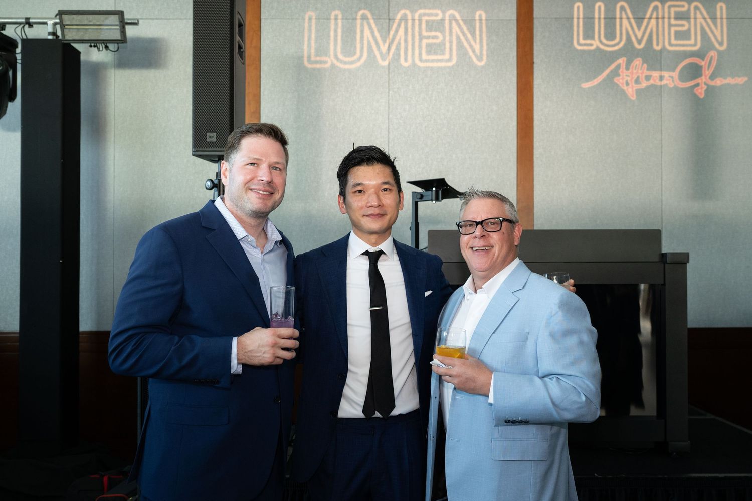 Three men smile and hold drinks at a Lumen After Dark event, dressed in suits at an upscale venue.