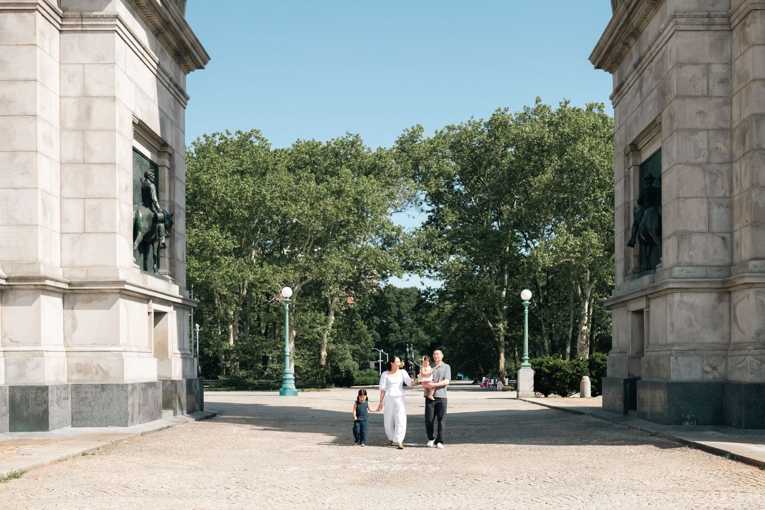 Family walking through grand stone archway entrance to a lush green park on a sunny day.
