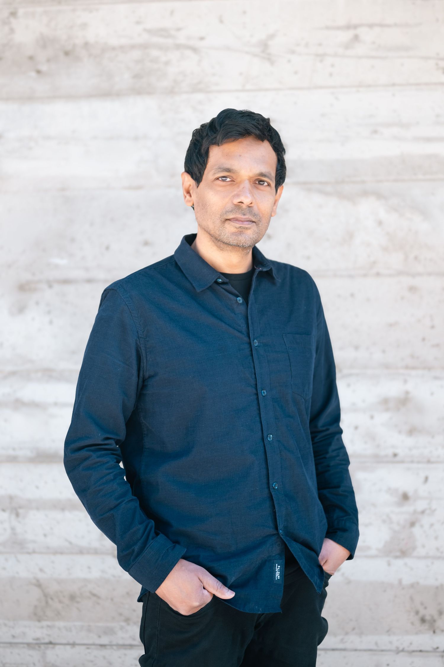 A man in a navy blue button-up shirt stands confidently with hands in pockets against a light stone wall.