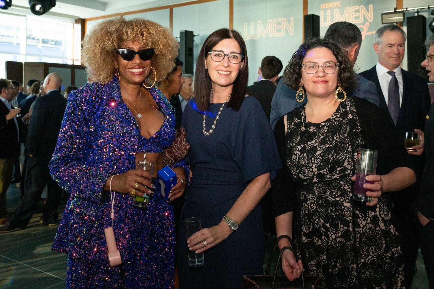 Three women pose together at an indoor gala event, one wearing a sparkling blue sequin outfit.