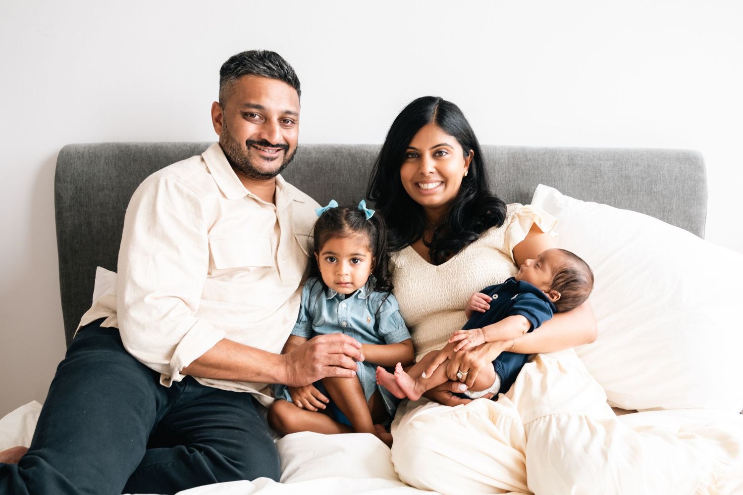 Happy South Asian family of four posing on a bed, parents smiling with two young children, cozy home setting.