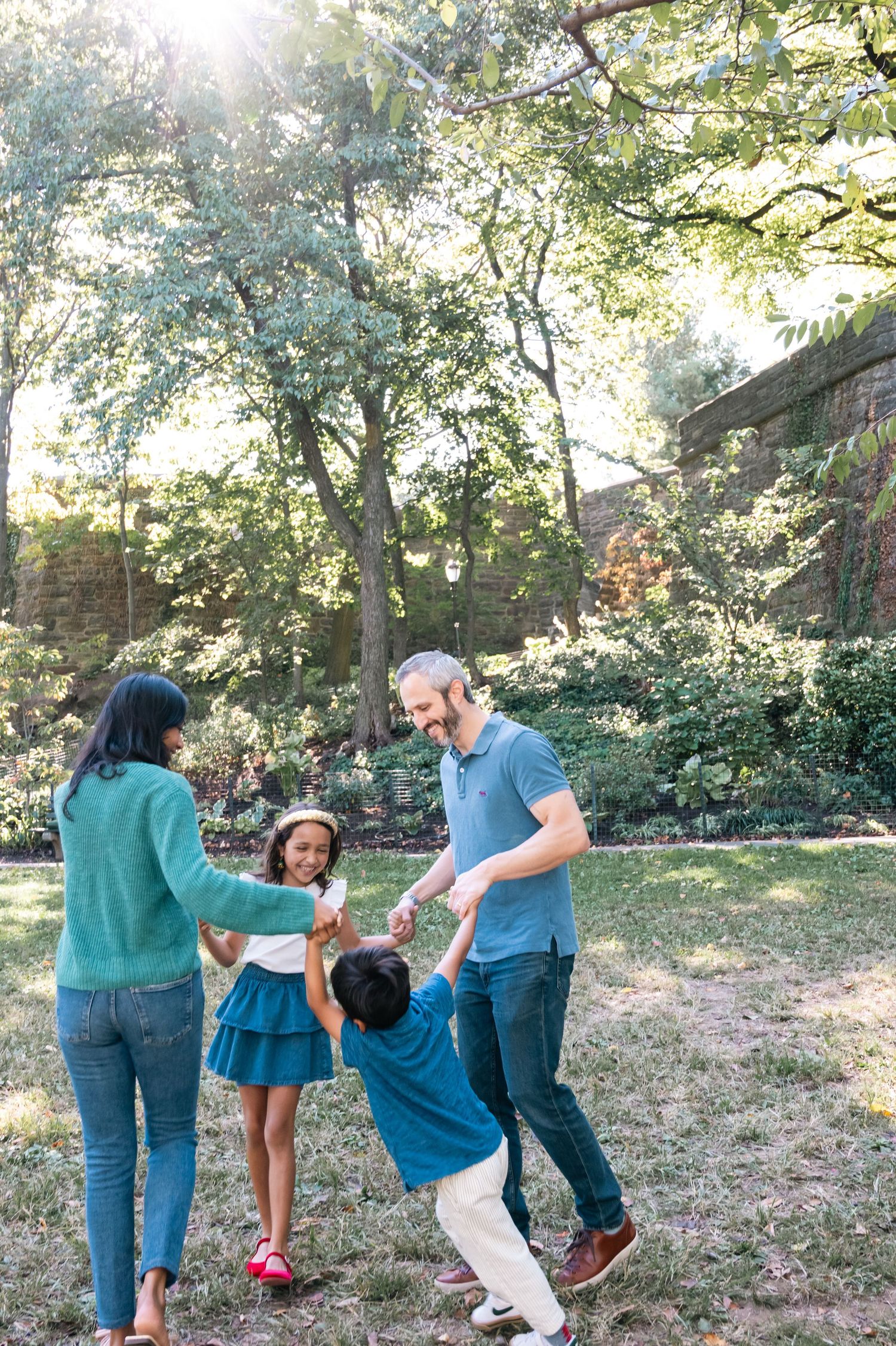 A family plays and dances together in a sunlit backyard garden surrounded by lush green trees.