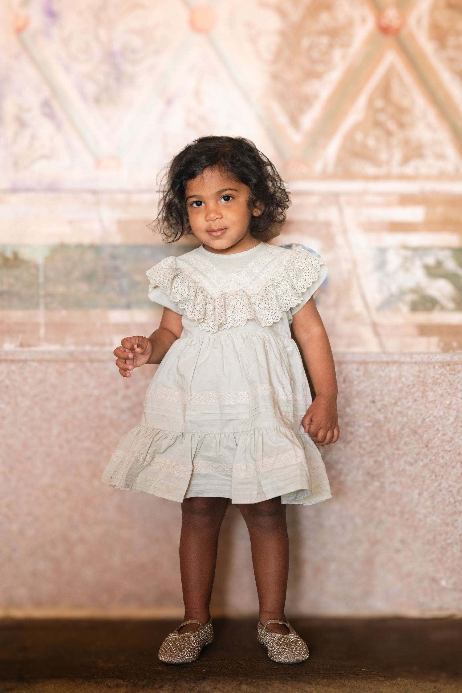 Toddler girl in white ruffle dress and gold shoes standing against a decorative pink wall.