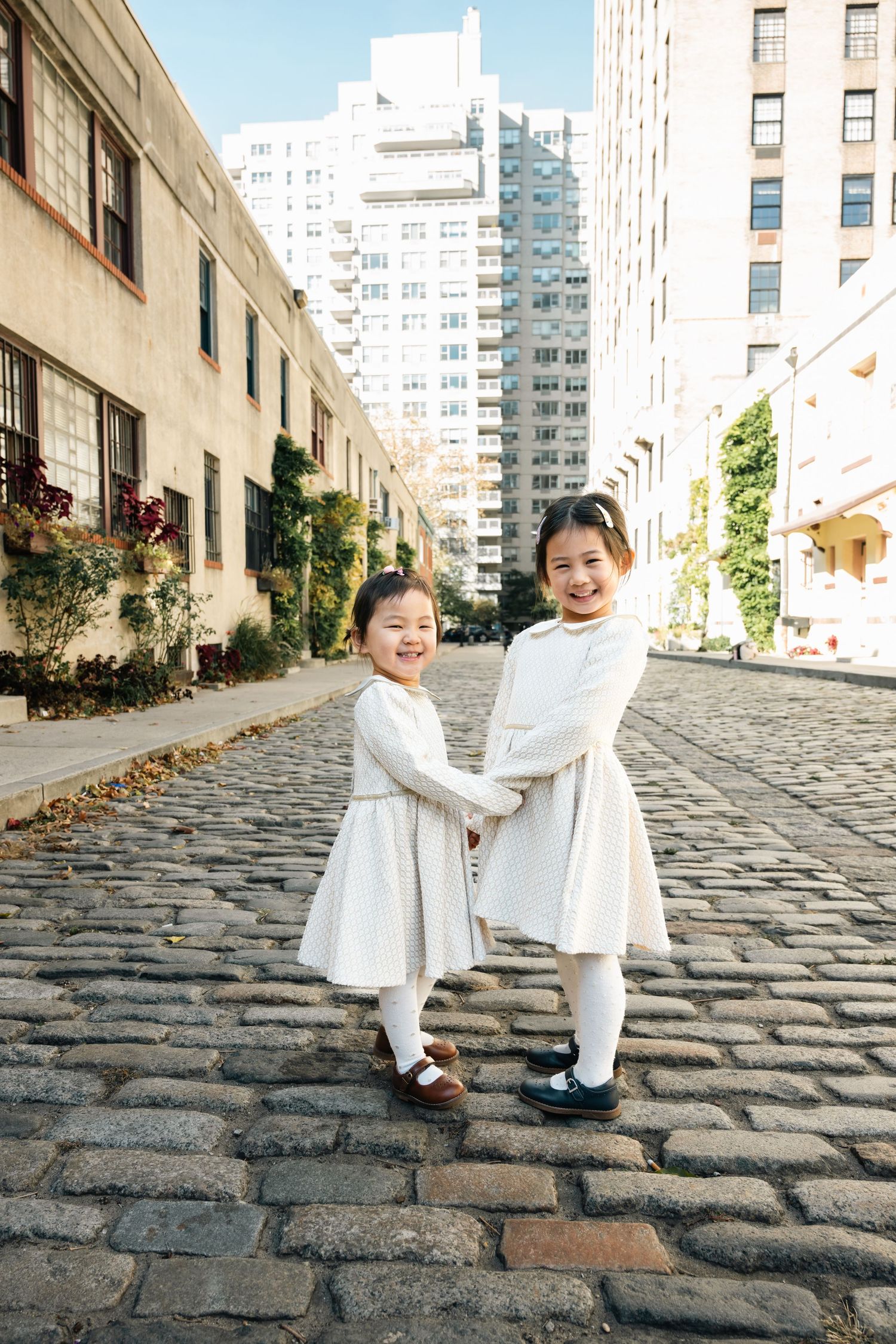 Two small figures in matching white outfits interact playfully on a cobblestone street.