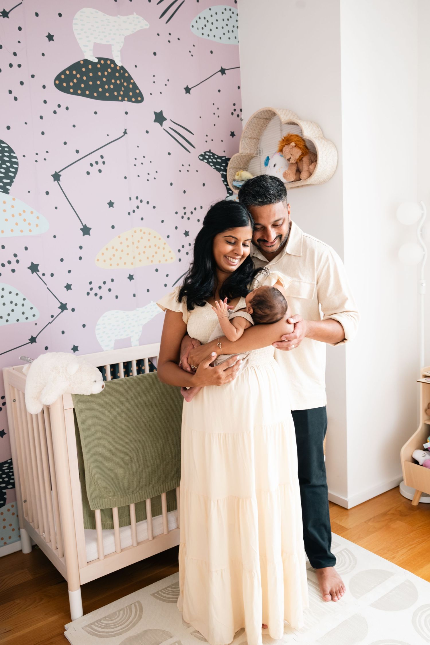 Nursery room features constellation wall art and white crib with parents holding newborn.