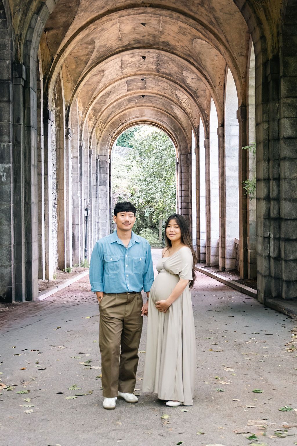 Couple poses under stone archway, pregnant woman in beige gown holds belly beside man in blue shirt.