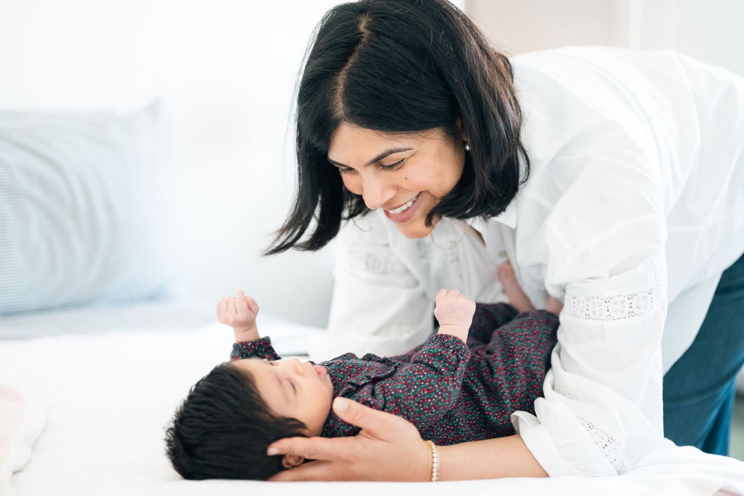 Smiling mother leaning over her newborn baby lying on a white bed, sharing a tender moment.