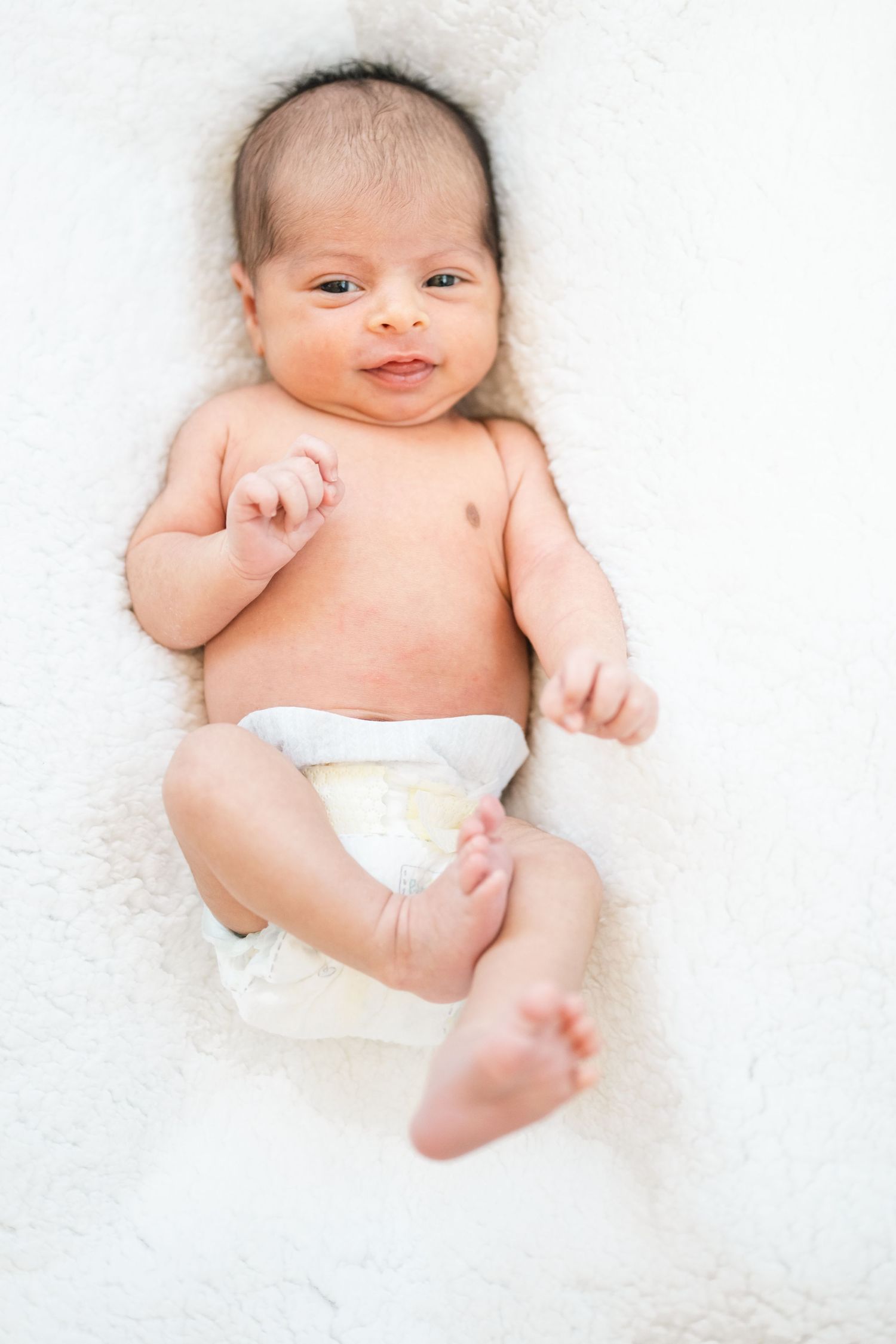 Newborn baby wearing white diaper lying on soft white blanket.