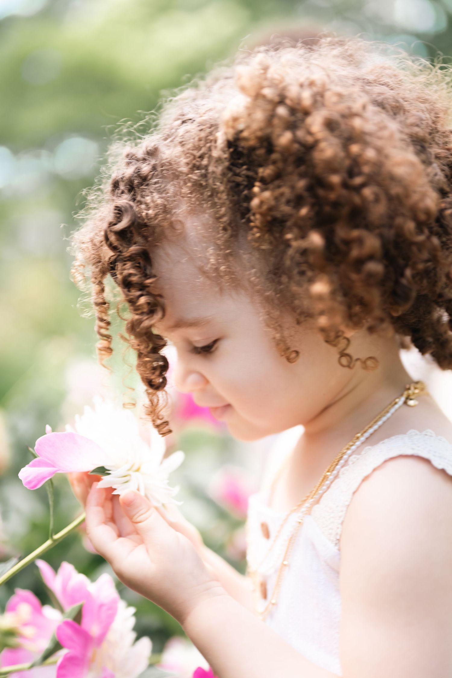 Curly-haired little girl smelling pink flowers in a sunny garden, wearing a white dress and gold necklace.