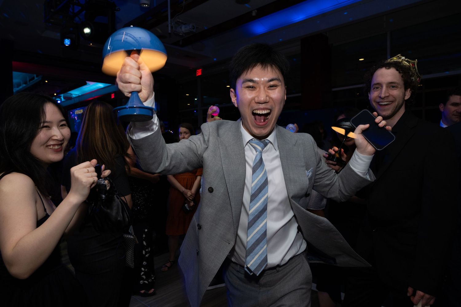 Excited man in gray suit celebrates winning a trophy at an awards event with friends cheering beside him.