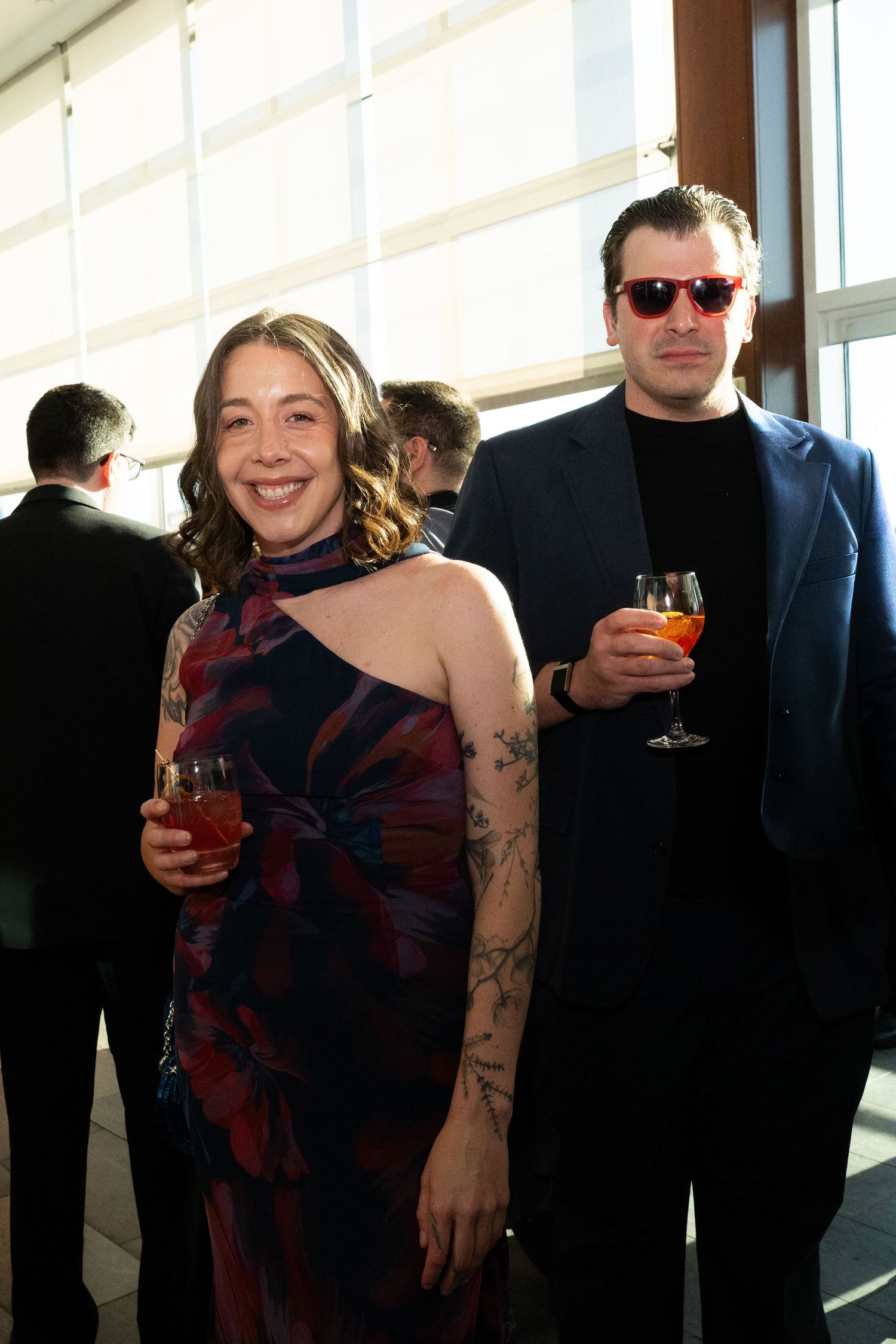 A smiling woman with tattoos in a floral dress holds a cocktail beside a man in sunglasses at a social event.