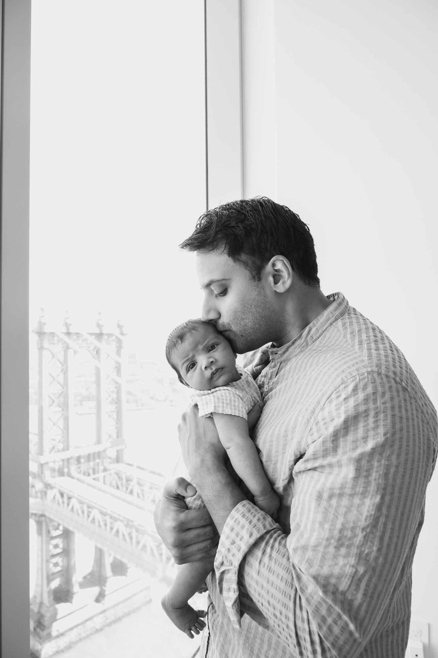 A tender moment between a parent and newborn baby against a window with a bridge visible in the background.