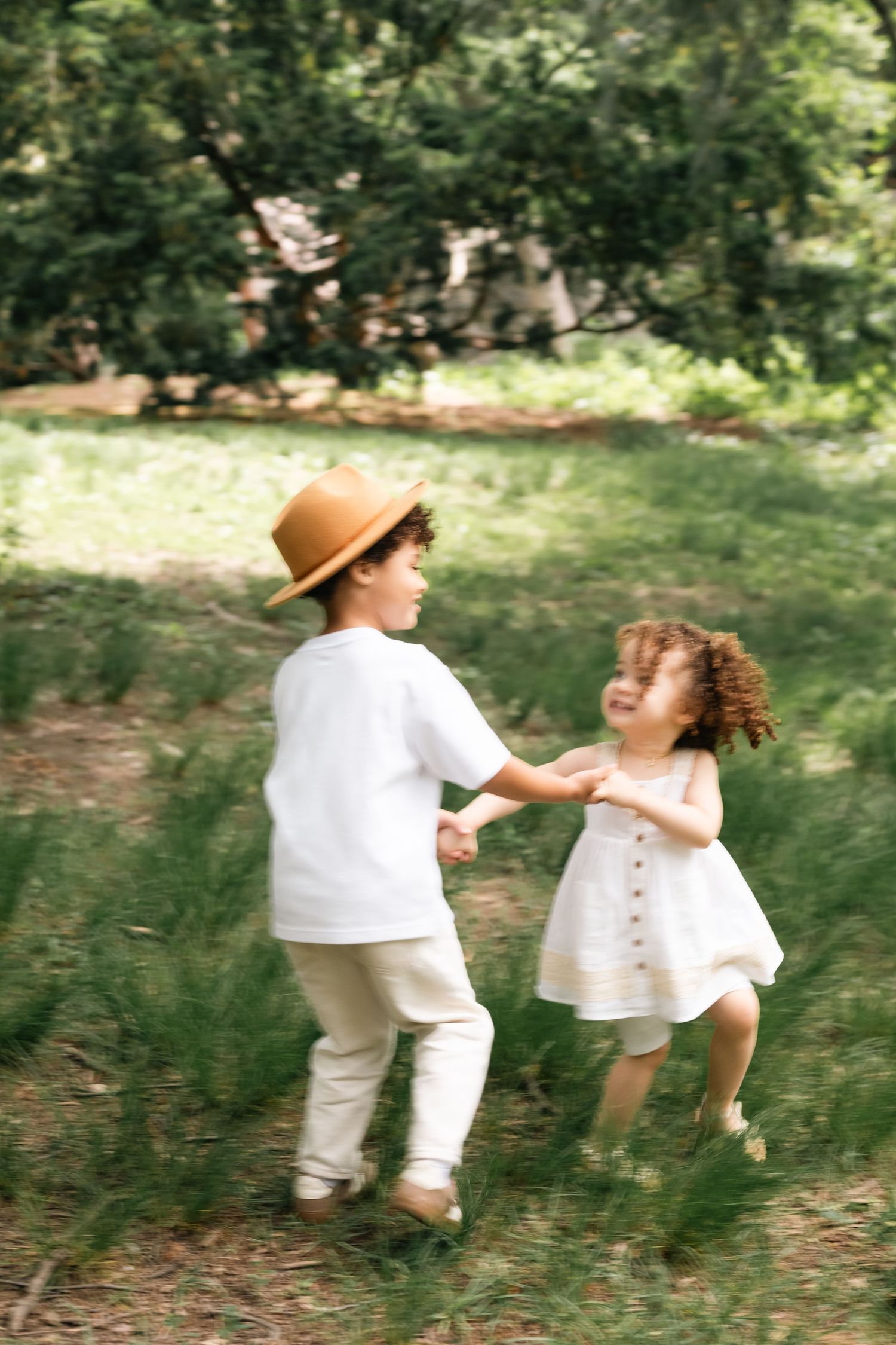 Two young children in white outfits holding hands and playing together on green grass in a sunny park.
