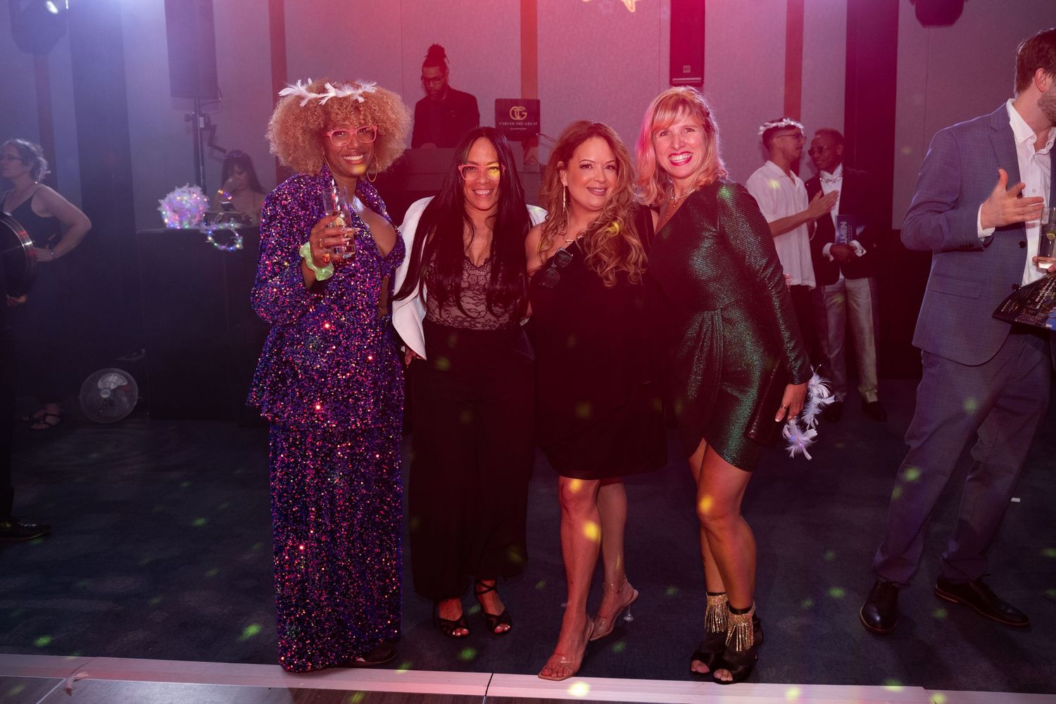 Four women in glamorous attire pose together smiling on a festive gala dance floor.