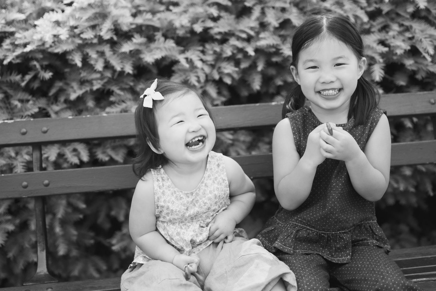 Two young Asian girls laugh joyfully while sitting on a bench in a black and white outdoor portrait.