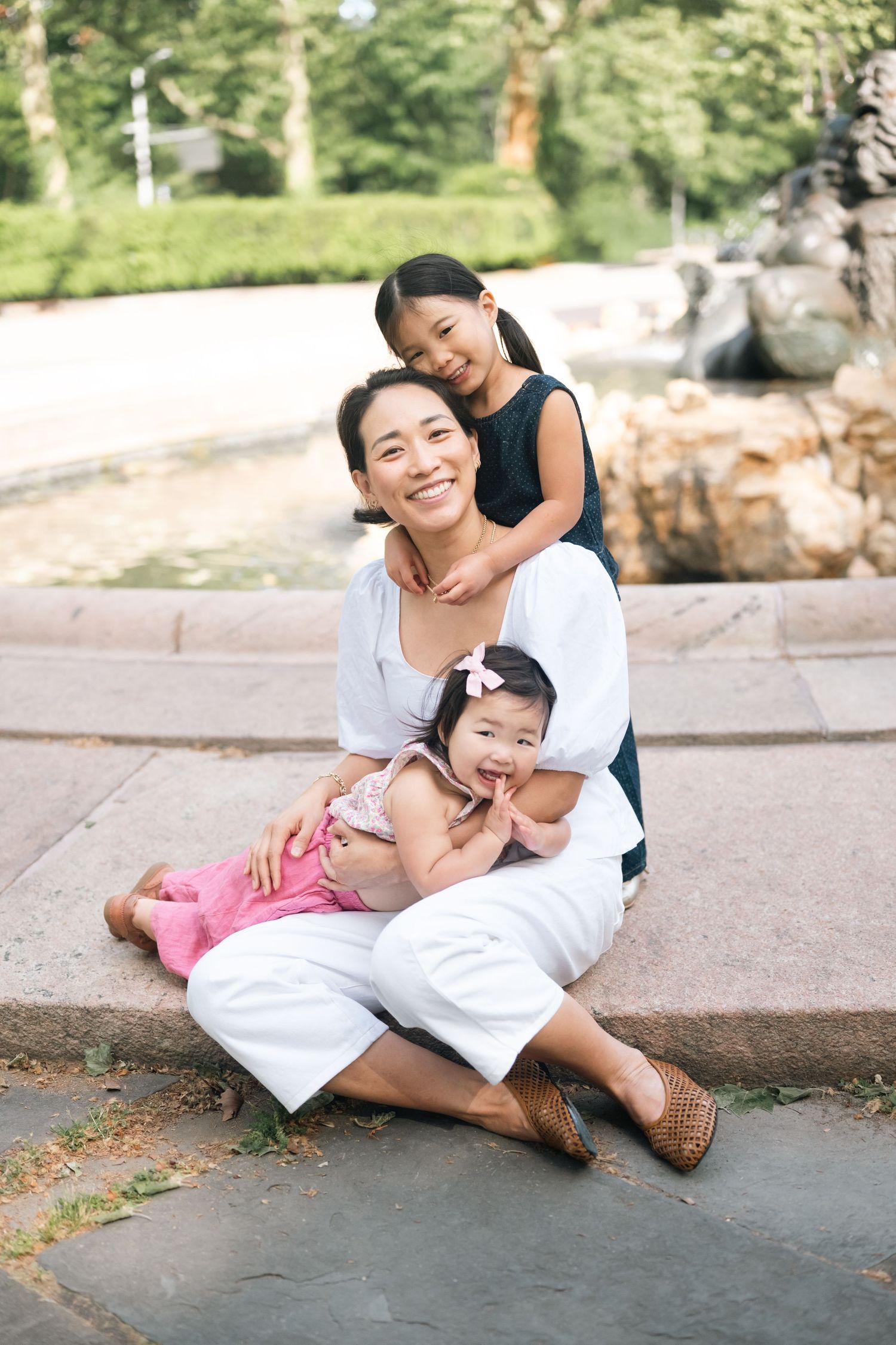 Smiling Asian mother sitting outdoors with two young daughters near a fountain, enjoying a warm family moment together.