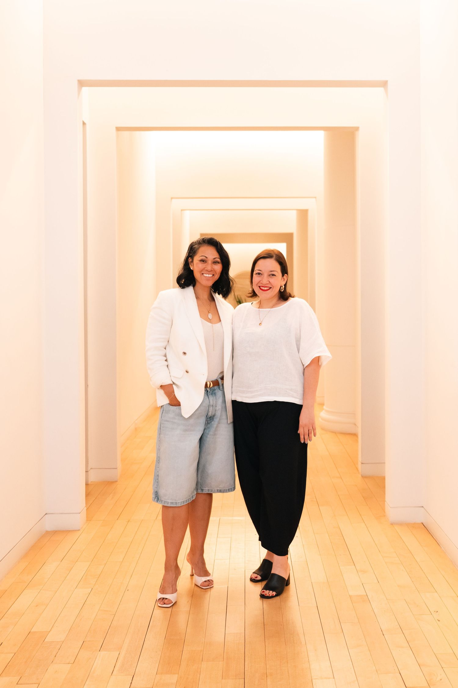 Two women in white tops smile together in a bright hallway with hardwood floors.