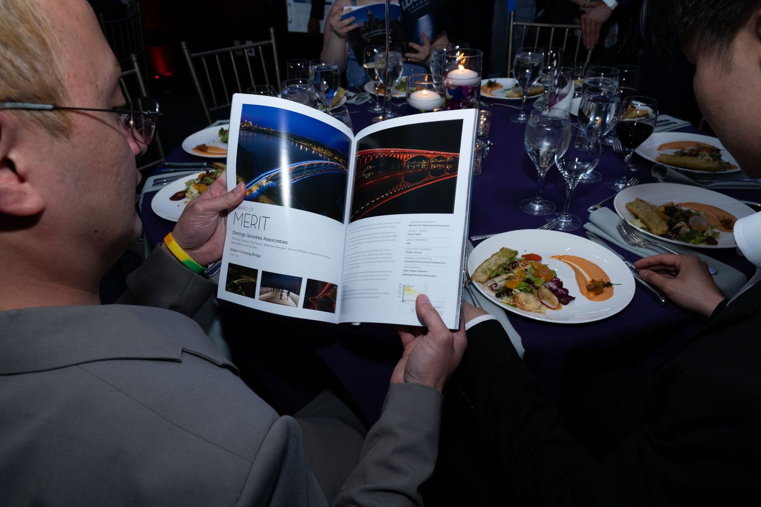 Man reading a glossy magazine at a formal dinner table with blue tablecloth and plated meals.