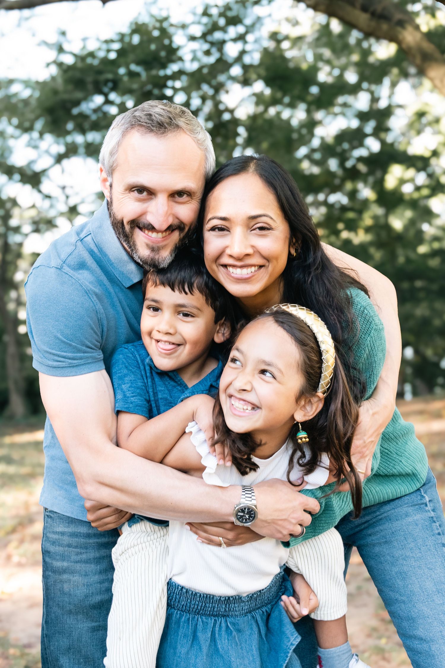 A happy family of four embraces and laughs together outdoors during a casual photo session in a park.