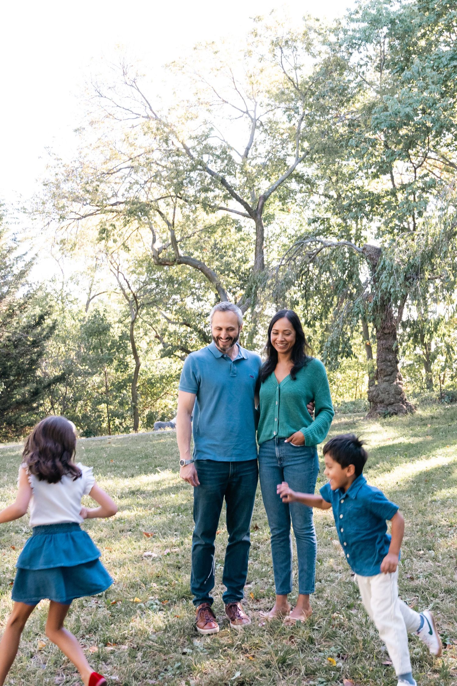 A family shares a playful moment outdoors on a grassy hill with trees in the background on a sunny day.