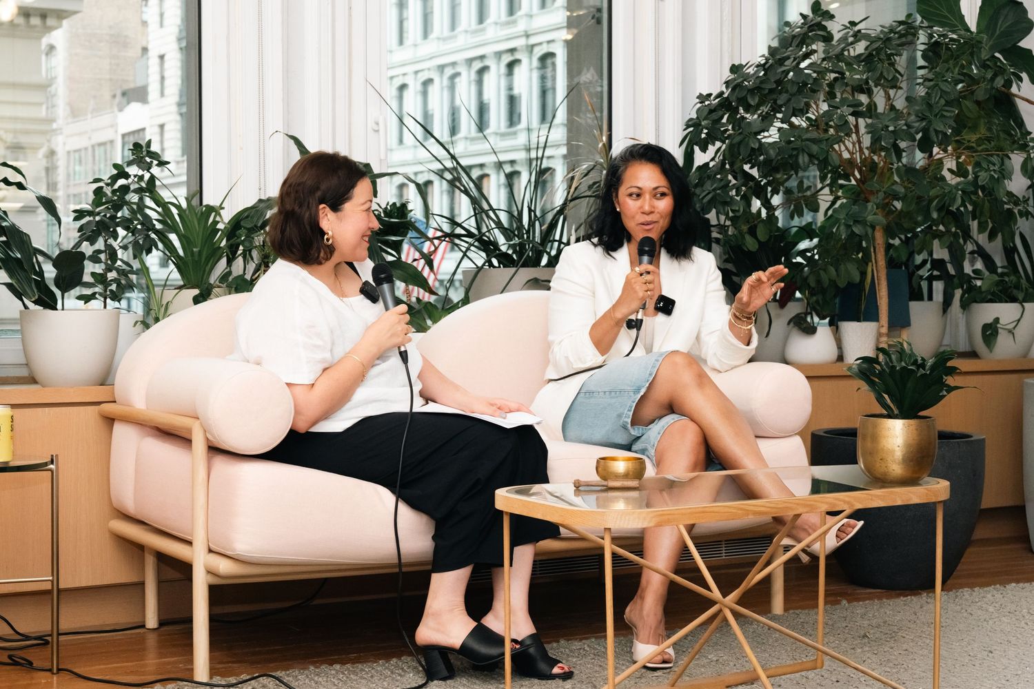 Two women sit on blush chairs holding microphones in a bright, plant-filled modern office event space.