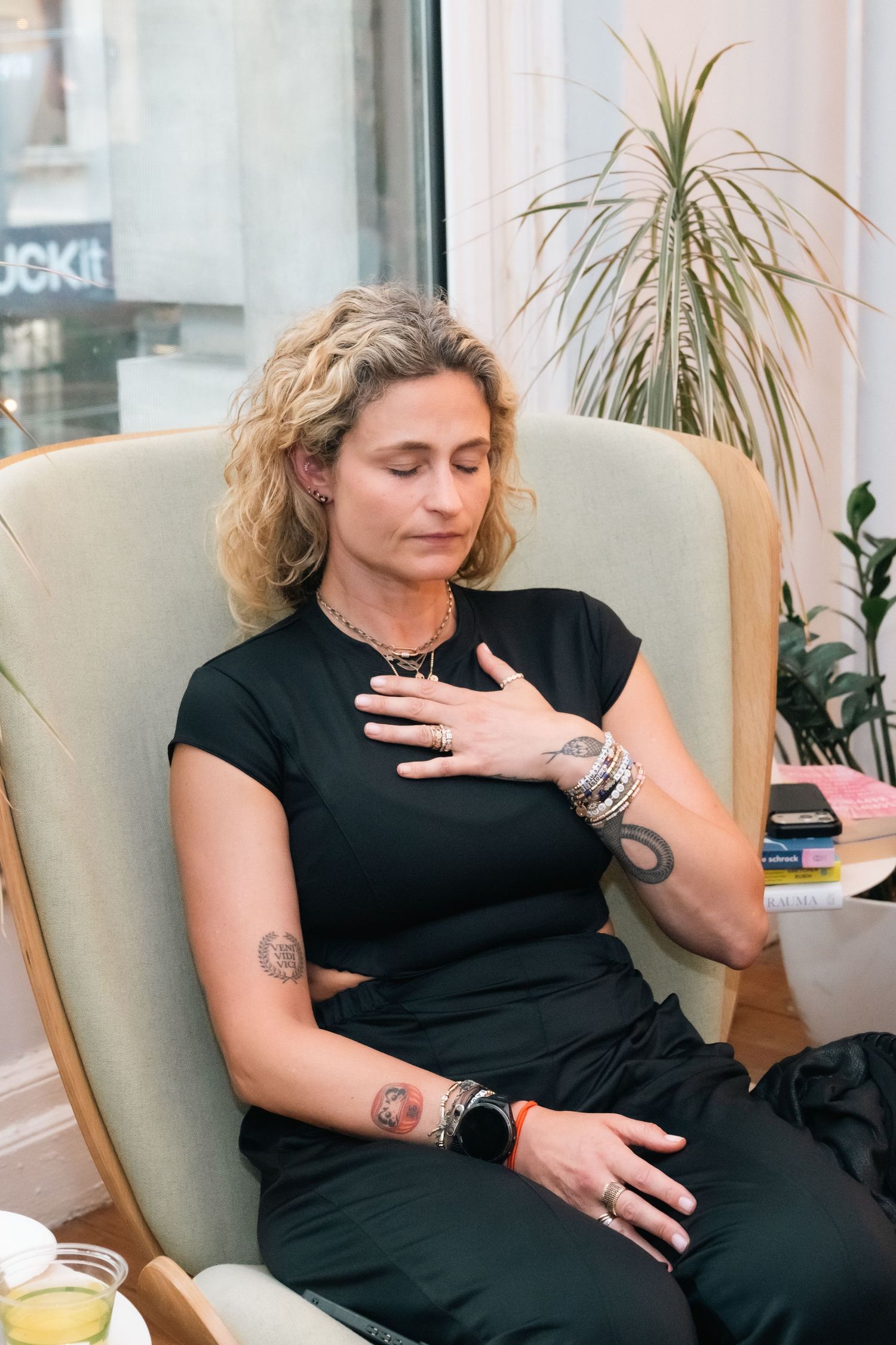 Woman in black outfit meditating in armchair with hand on chest, tattoos visible, eyes closed, near window.