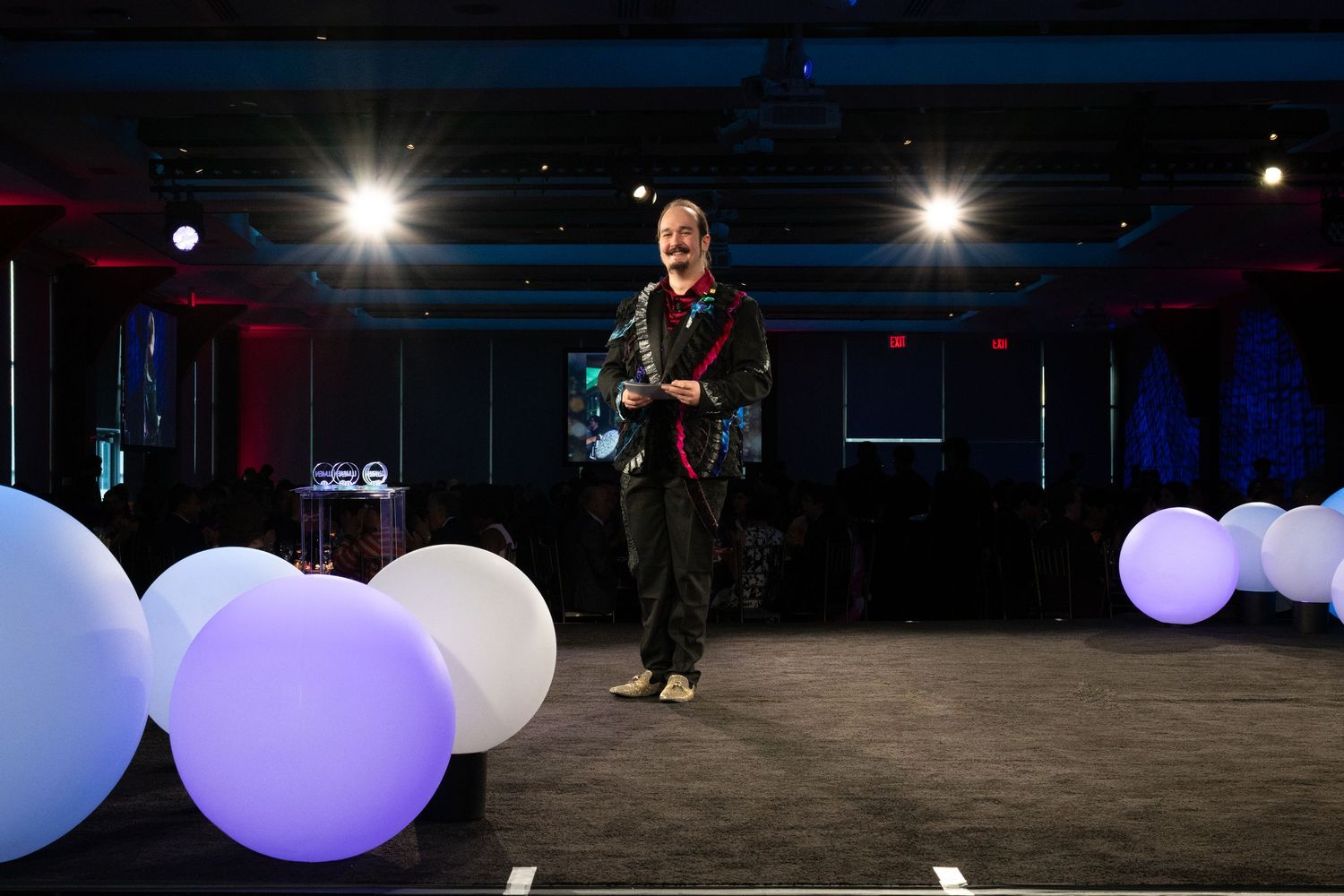 Performer in colorful outfit stands on stage surrounded by glowing white spherical light props.