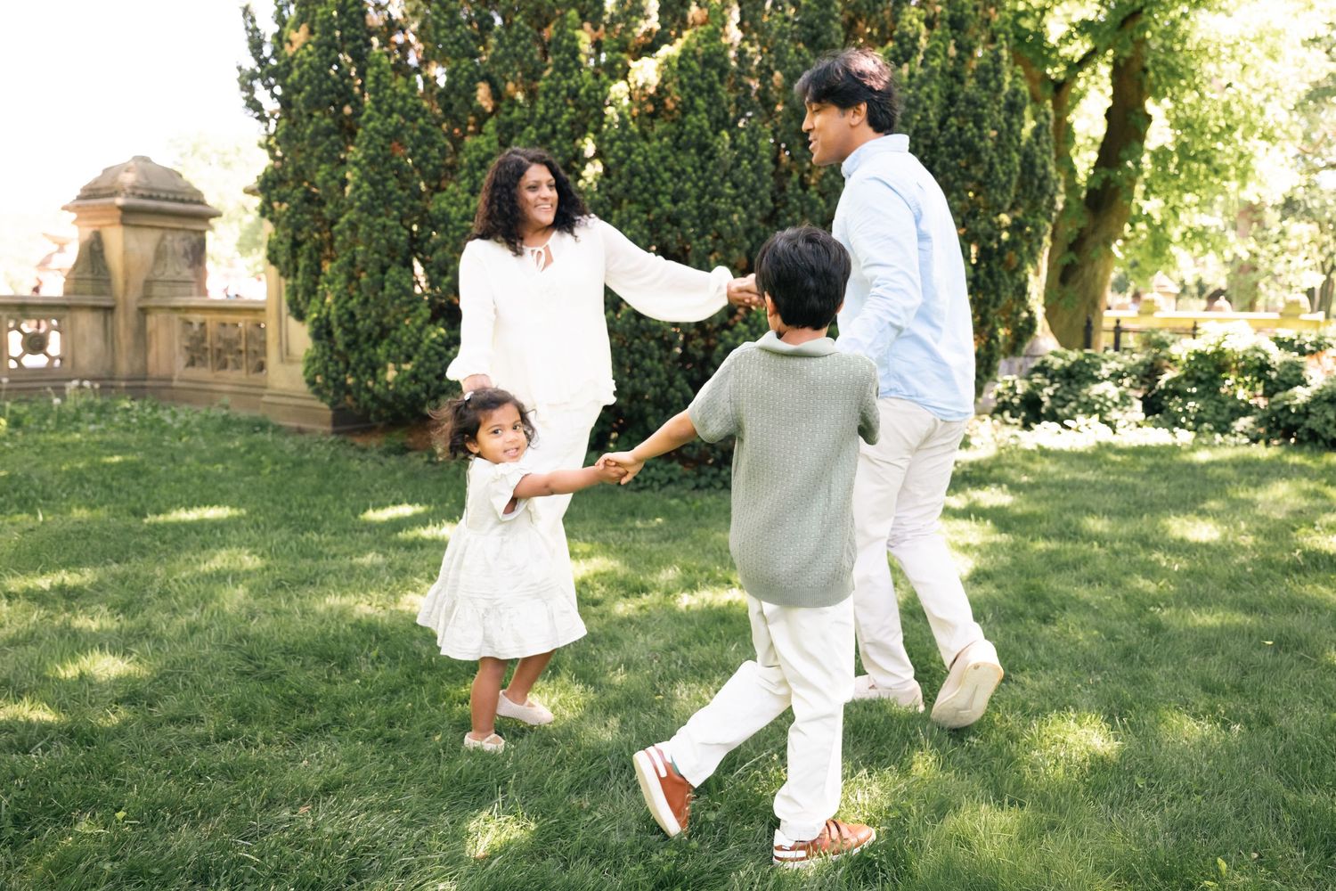 Family of four holding hands and dancing together on green grass in a sunny park setting.