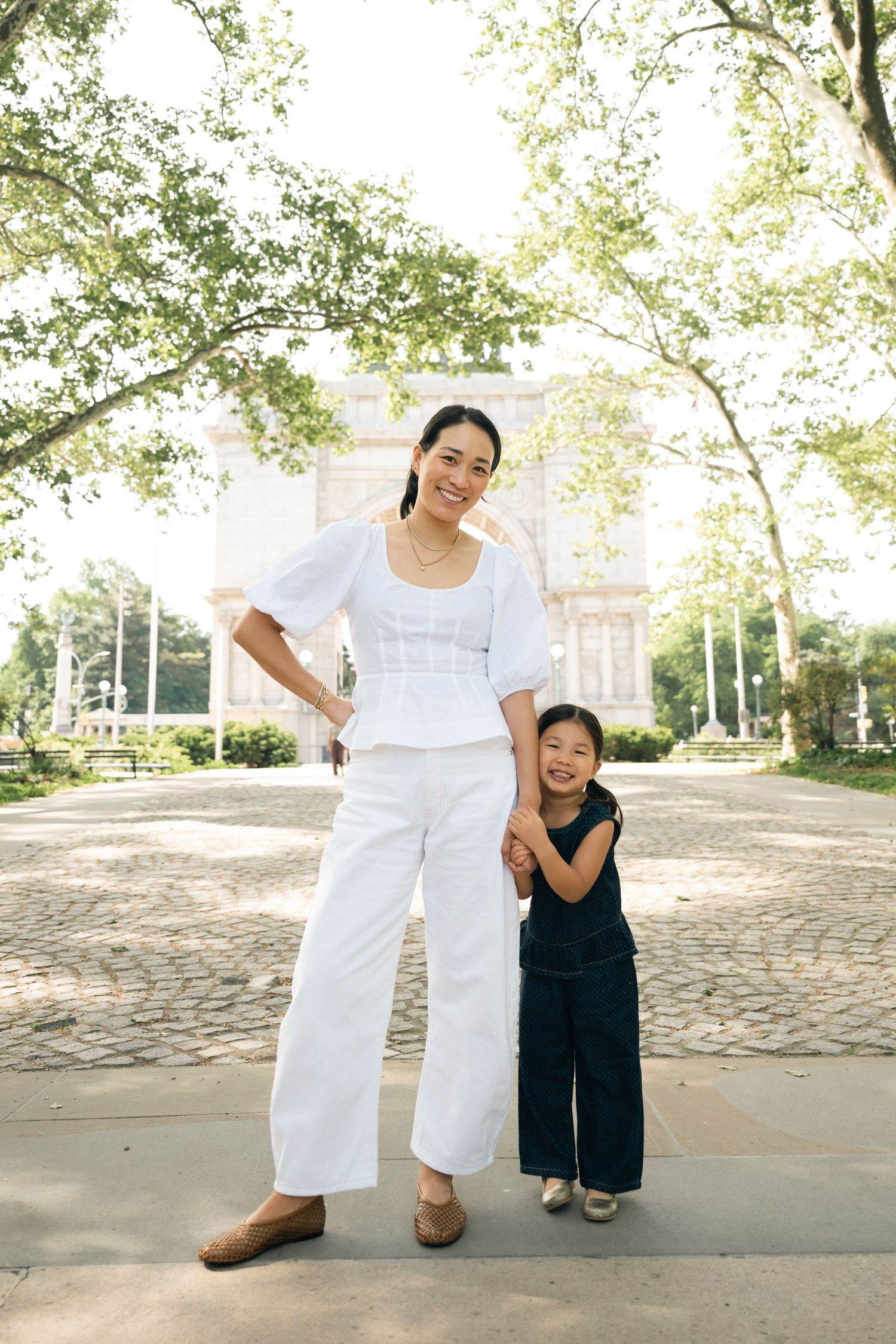 A woman in white outfit holds hands with a young girl in black in a sunny park setting.