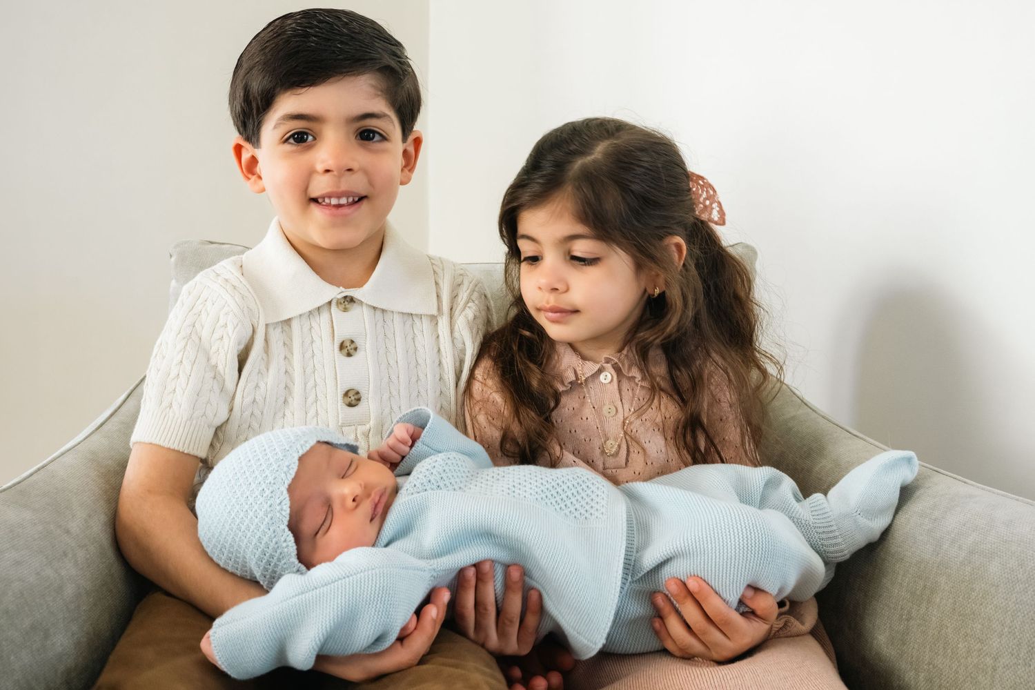 Family members gathered on grey sofa sharing tender moments with newborn in light blue blanket.