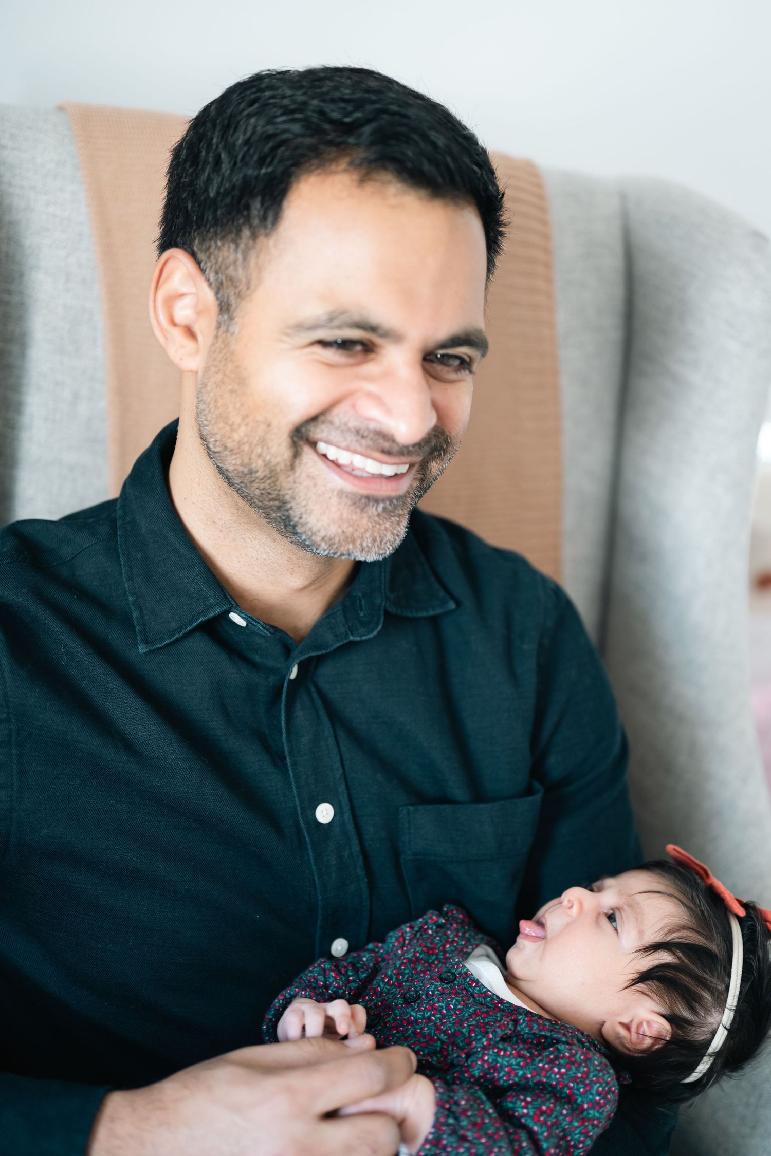 Smiling man with short dark hair and beard holding a sleeping newborn baby girl in a red headband.