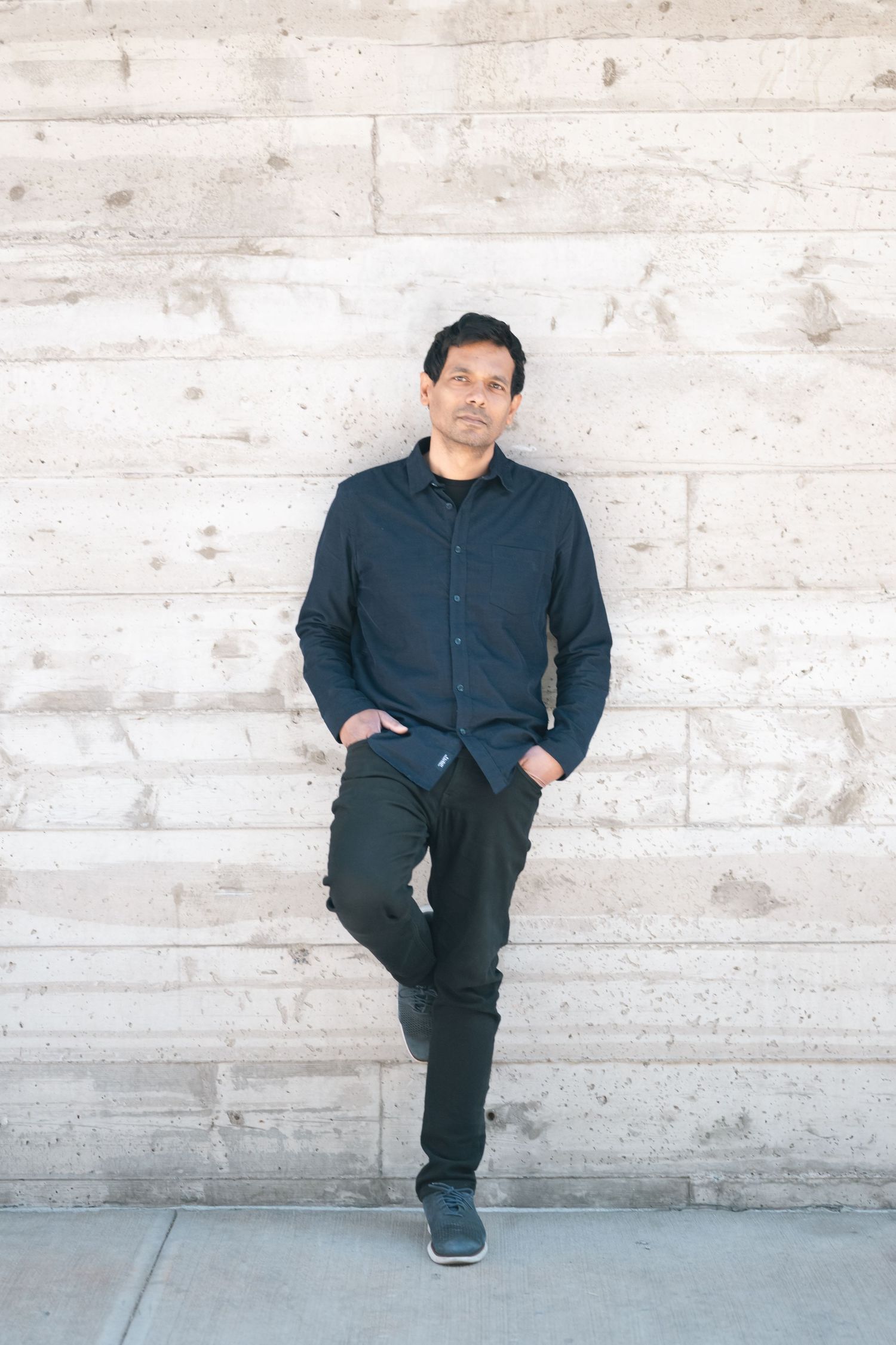 A man in a dark blue shirt and black pants leans against a concrete wall with hands in pockets.