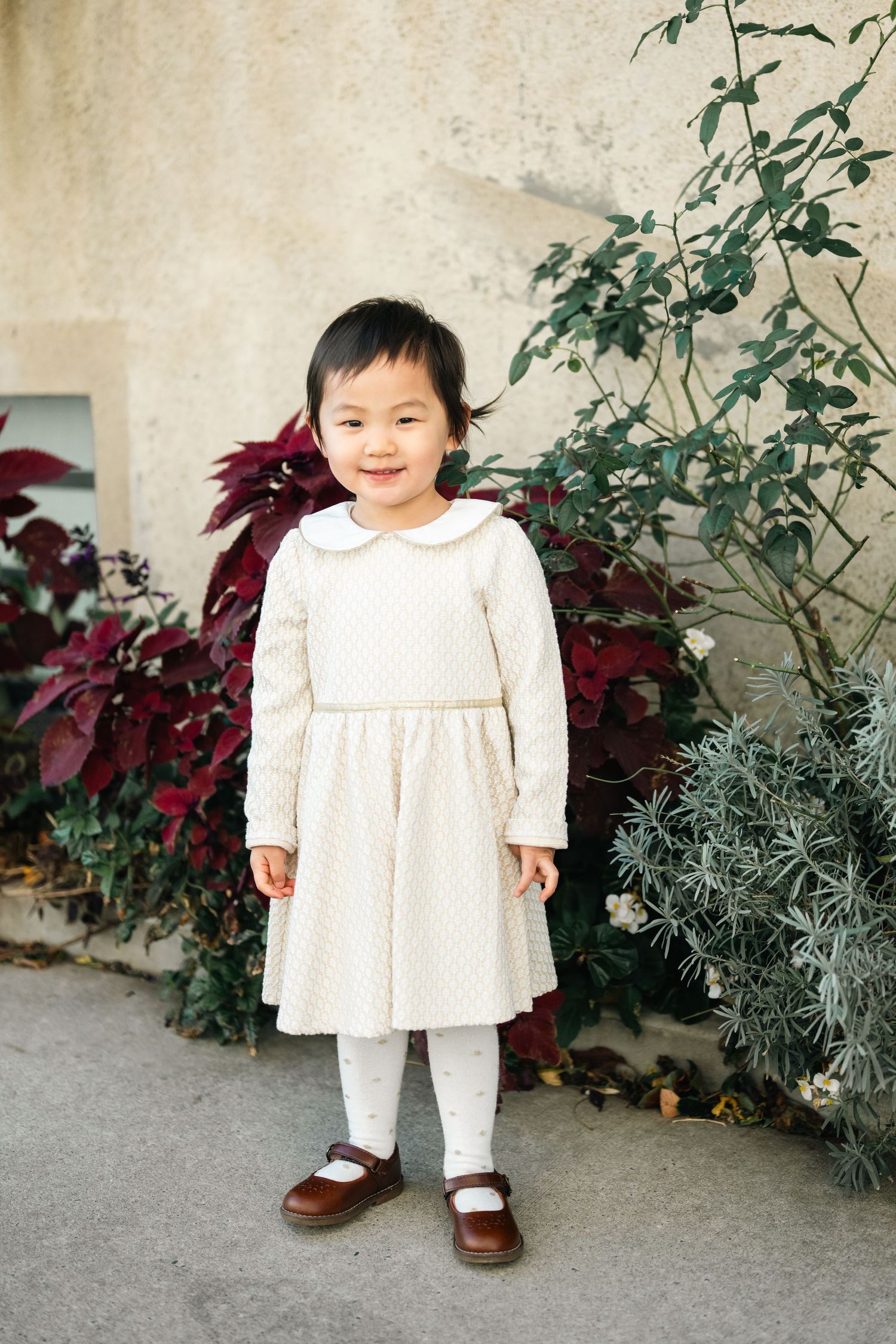 A small child in a white dress stands near decorative plants and a stone wall.