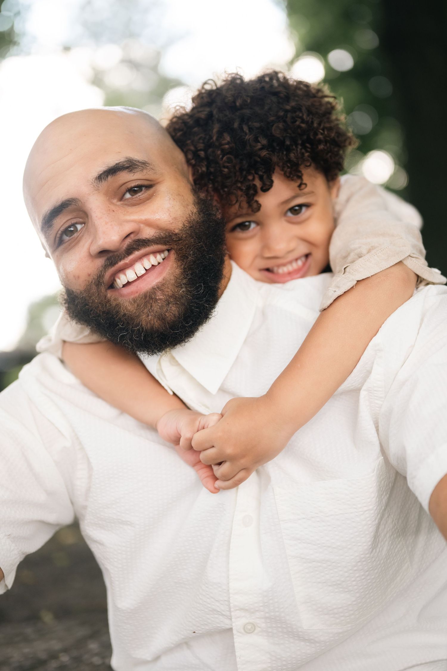 A bald bearded man smiles while a curly-haired child hugs him from behind outdoors.