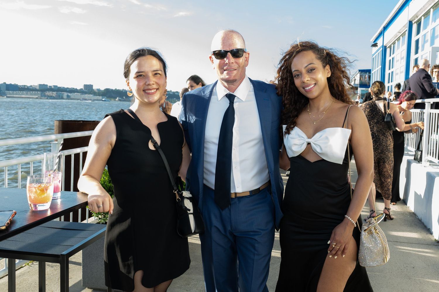 Three elegantly dressed guests posing together at an outdoor waterfront evening event with city views.