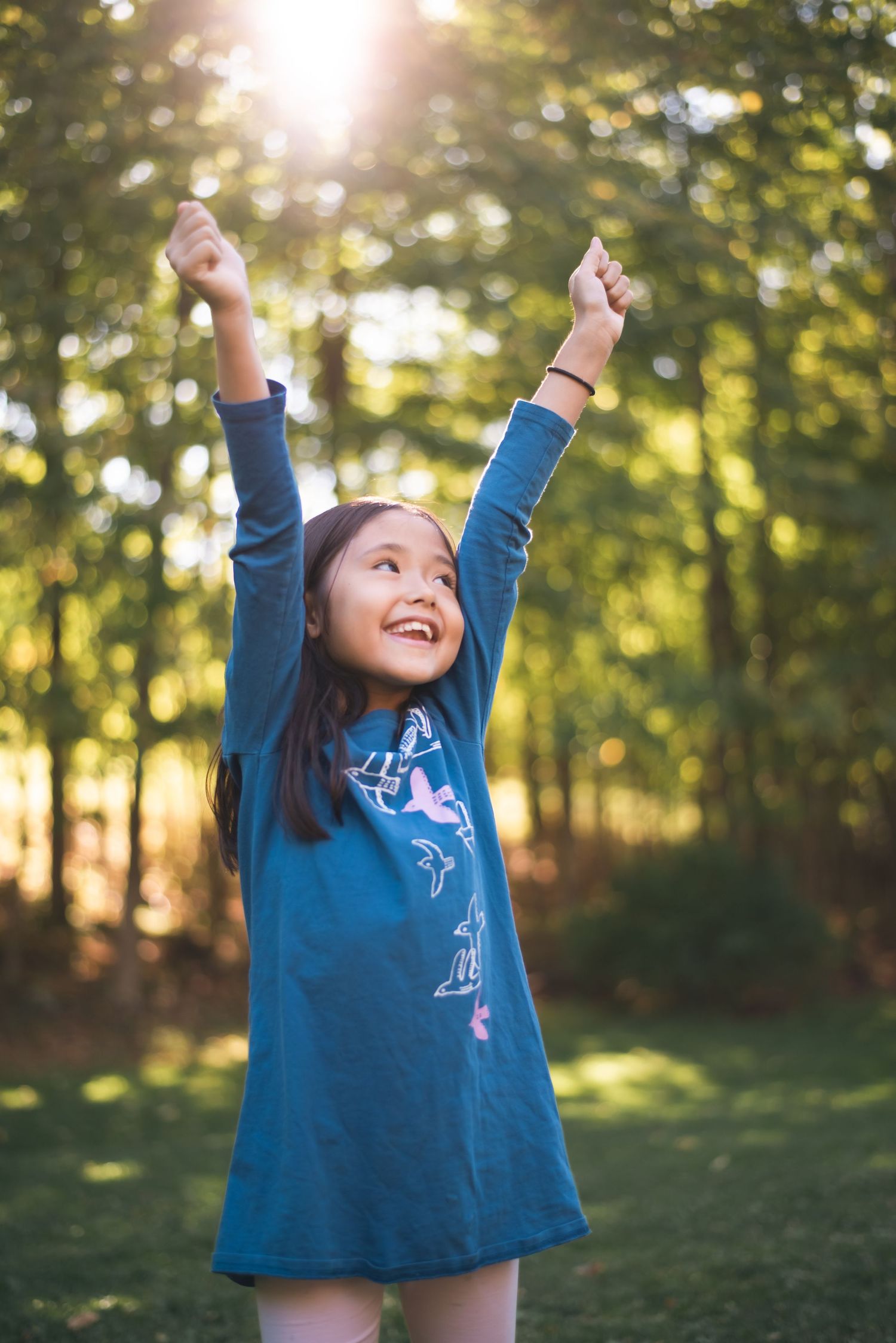 Child in blue dress reaching joyfully towards sunlit sky in garden.