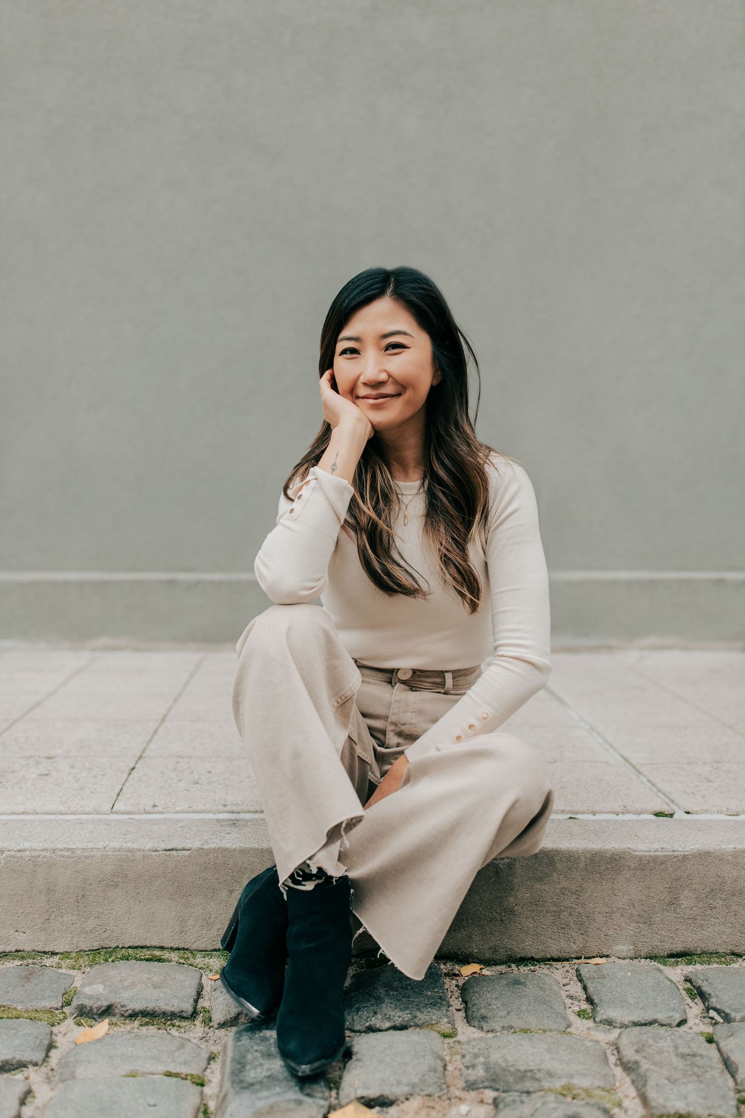 Person in beige coat sitting on sidewalk curb during outdoor photoshoot.
