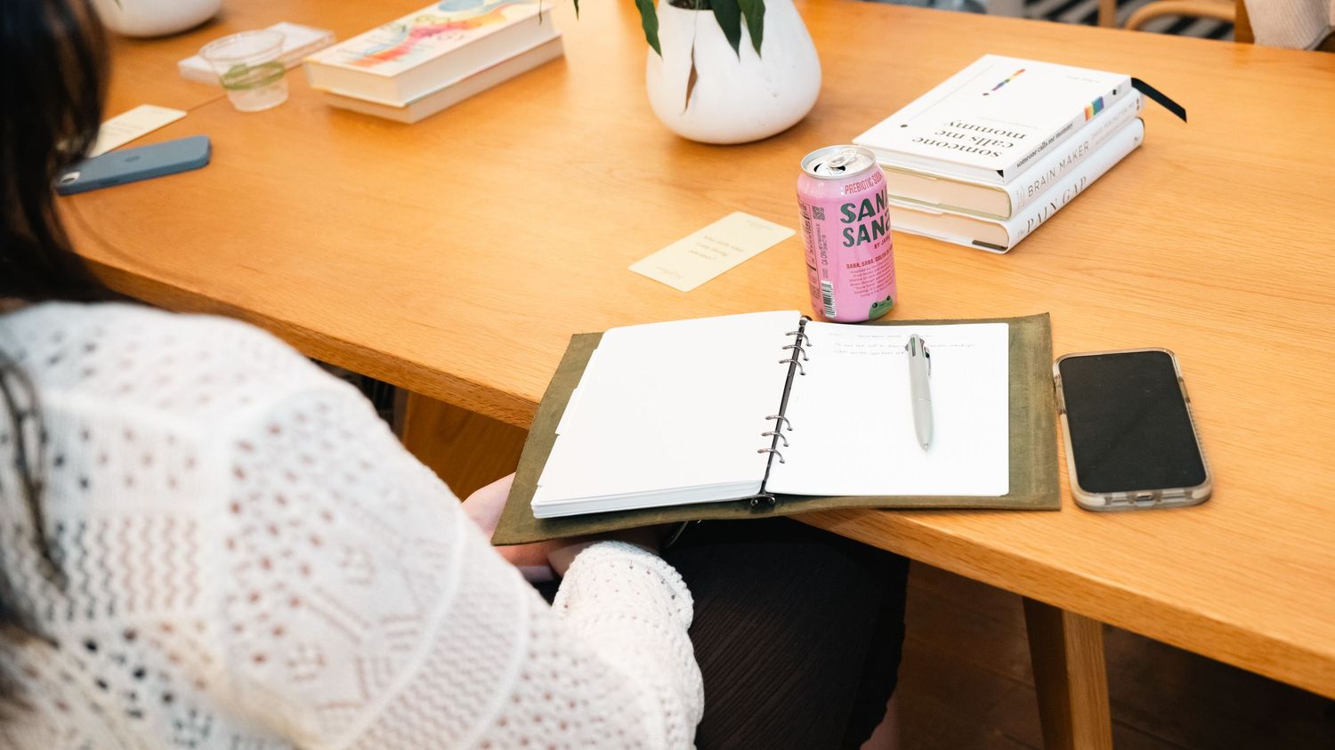 Woman in white sweater writing in open planner notebook at wooden desk with pink can and smartphone.