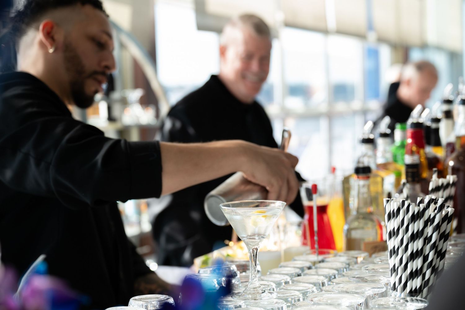 Bartender pouring a cocktail into a glass at a bar with bottles and drinks in the background.