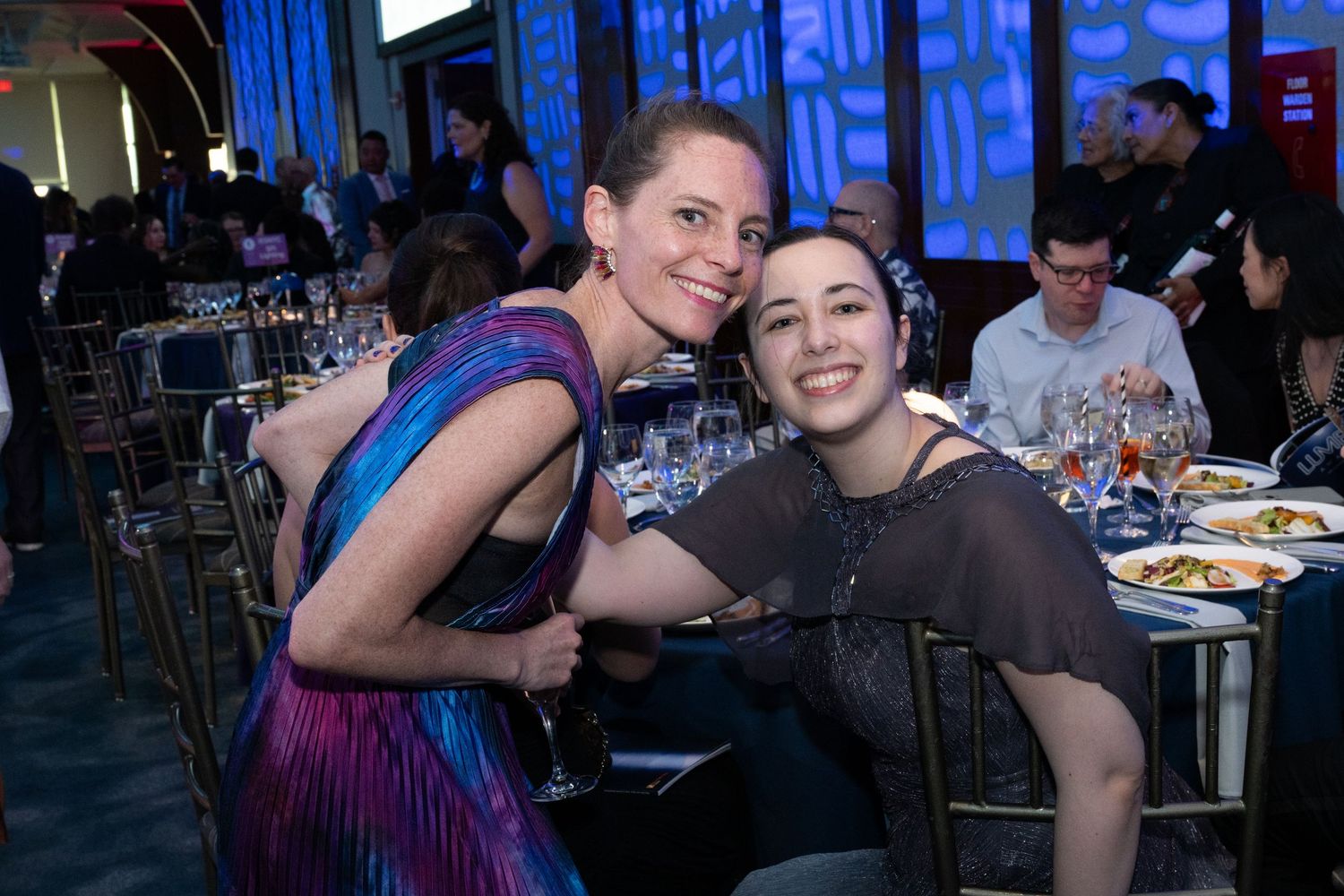 Two women smile and embrace warmly while seated at a formal banquet dinner event.