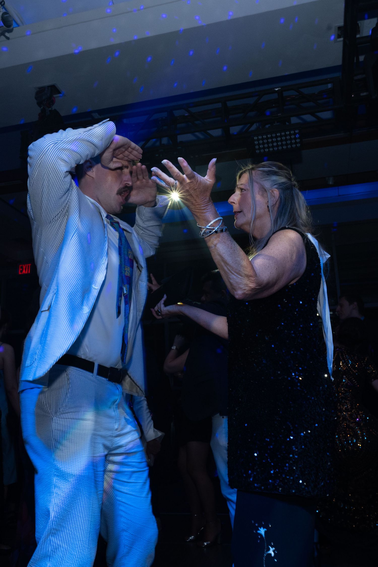 Two people dancing energetically at a nightclub party, man in white suit and woman in black sequin dress under blue lights.