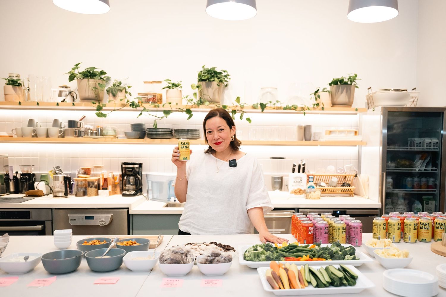 Woman in white top holds juice glass behind a counter with colorful bowls, vegetables, and bottled drinks in a bright kitchen.
