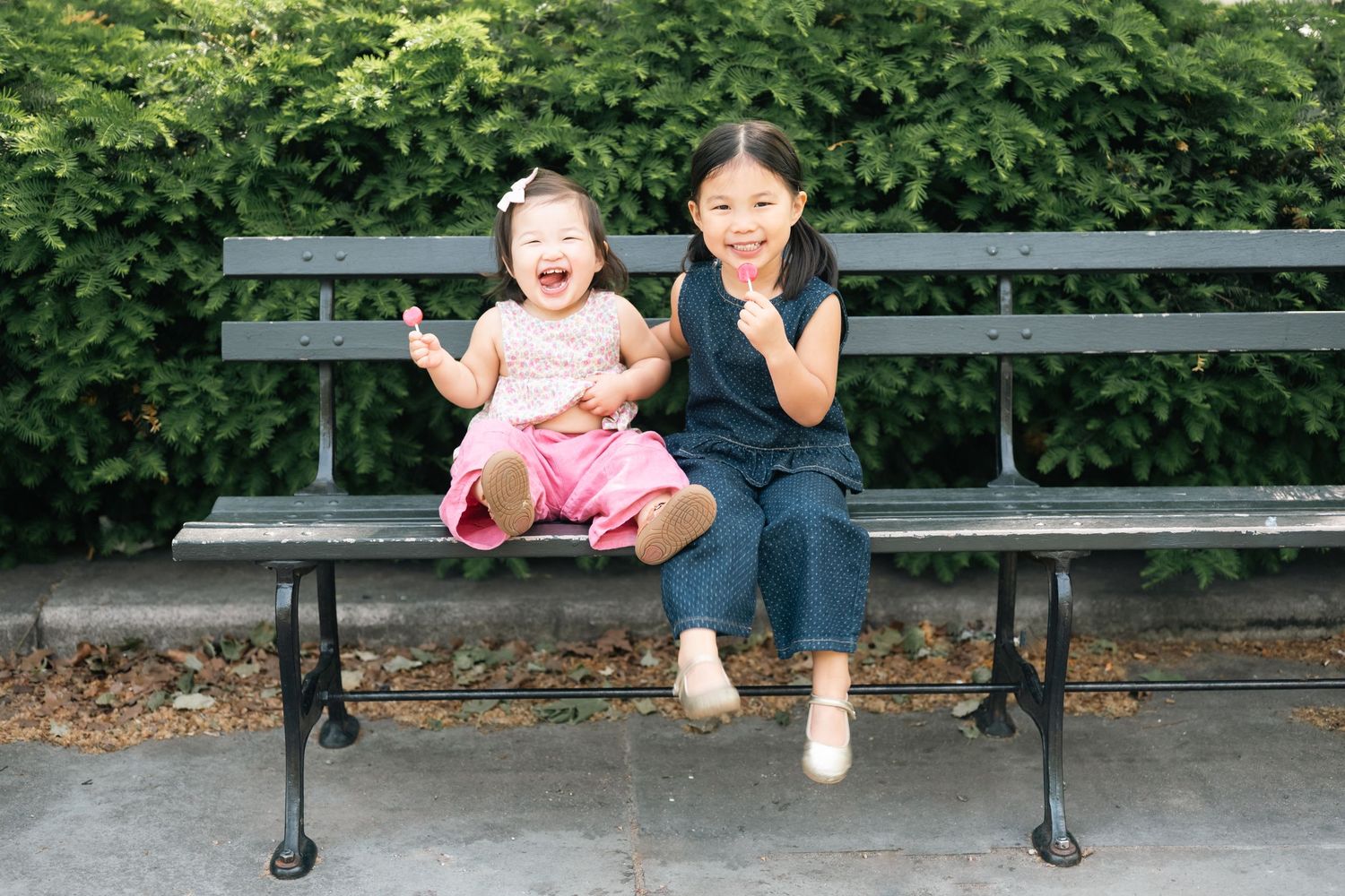 Two young Asian girls sitting on a park bench, smiling and laughing outdoors surrounded by green shrubs.