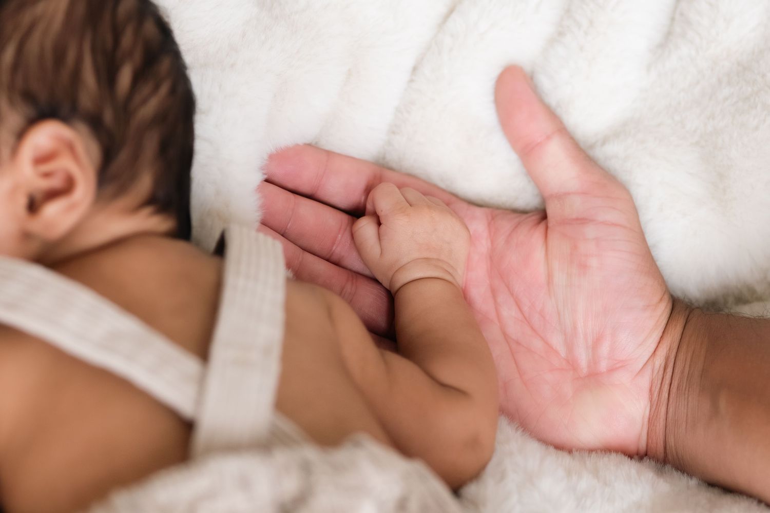 A small hand gently holds onto an dad's palm while resting on white bedding.
