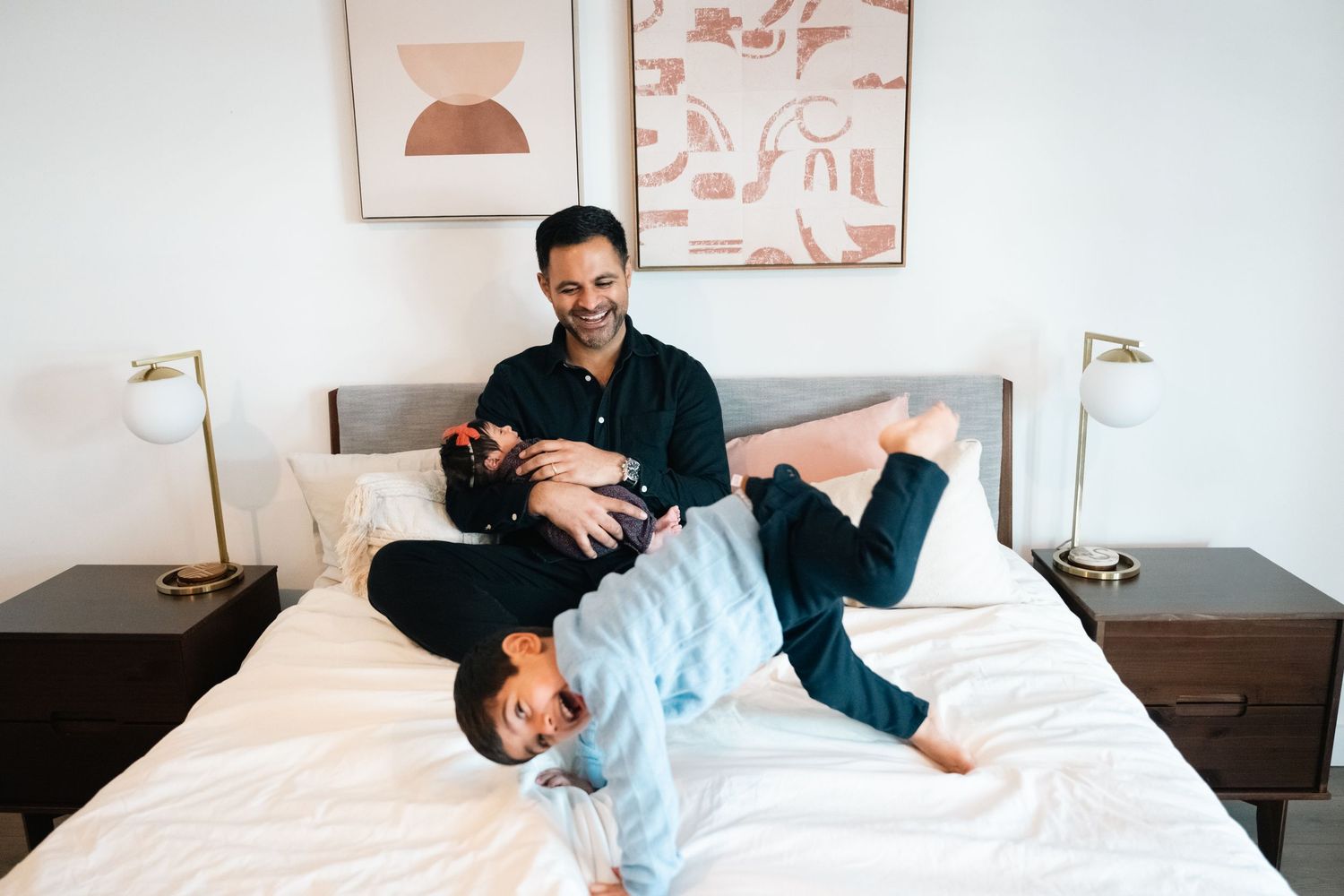 A smiling man plays with a laughing child on a white bed in a bright, modern bedroom with framed art.